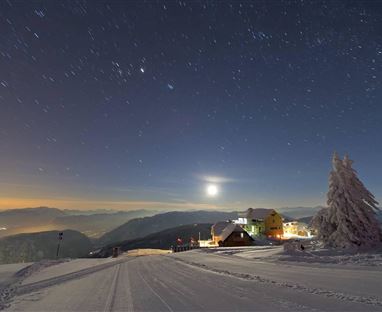 A winter landscape with snow-covered hills and a house. The sky is clear with many stars and a shining moon.