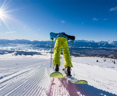 Ein Skifahrer fährt auf frisch präparierter Piste. Im Hintergrund sind schneebedeckte Berge und ein klarer, blauer Himmel zu sehen.