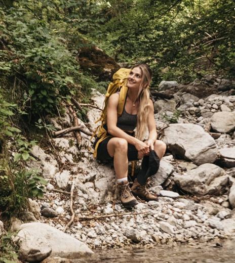 Eine Frau sitzt auf Steinen neben einem klaren Bach in einem Wald. Sie trägt einen Rucksack und wirkt entspannt in der Natur.