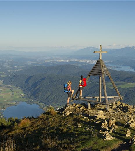 Zwei Wanderer stehen an einer Aussichtsplattform mit einem Holzkreuz auf einem Berggipfel. Im Hintergrund sind weite Täler und Berge mit klarem Himmel zu sehen.