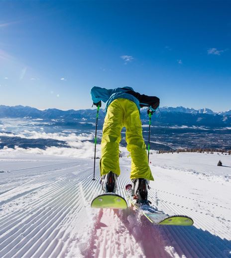 Ein Skifahrer fährt auf frisch präparierter Piste. Im Hintergrund sind schneebedeckte Berge und ein klarer, blauer Himmel zu sehen.