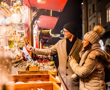 Ein Paar schaut sich die Waren an einem Weihnachtsmarkt an. Die Stände sind festlich beleuchtet und die Atmosphäre ist gemütlich.