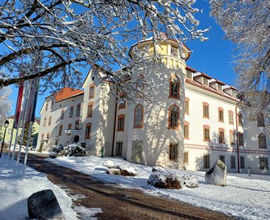 Ein schönes, historisches Gebäude im Schnee. Die Umgebung ist winterlich mit schneebedeckten Bäumen und einem blauen Himmel.