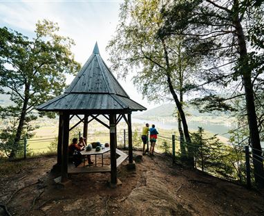 Eine Holzpavillon umgeben von Bäumen mit Blick auf den See. Zwei Personen stehen und genießen die Aussicht.