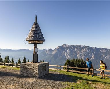 Eine Wandergruppe auf einem Berg mit Blick auf majestätische Berge. Im Vordergrund steht ein kleiner Aussichtspunkt mit einem geschützten Dach.