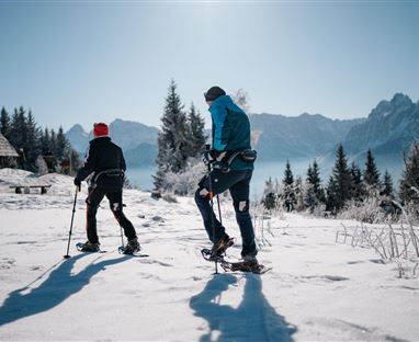 Zwei Personen wandern im Schnee mit Schneeschuhen. Im Hintergrund sind Berge und ein klarer blauer Himmel zu sehen.