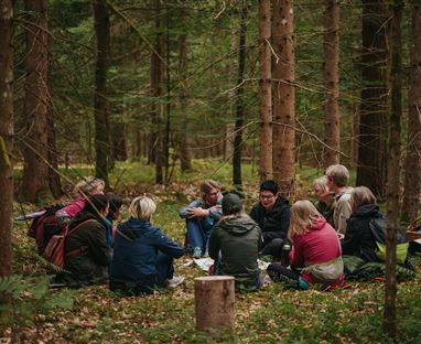 Eine Gruppe von Menschen sitzt im Wald und spricht miteinander. Um sie herum wachsen Bäume und das Laub liegt auf dem Boden.