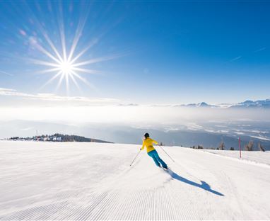 Ein Skifahrer fährt die Piste hinunter. Im Hintergrund ist ein klarer blauer Himmel und die Sonne strahlt über die schneebedeckte Landschaft.
