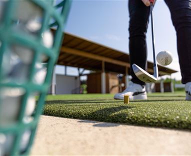 Eine Person spielt Golf und hat gerade den Ball abgeschlagen. Im Hintergrund sind ein Golfplatz und eine Überdachung zu sehen.