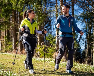 Ein Paar wandert mit Nordic-Walking-Stöcken auf einem schmalen Weg im Wald. Die Sonne scheint und der Himmel ist klar.