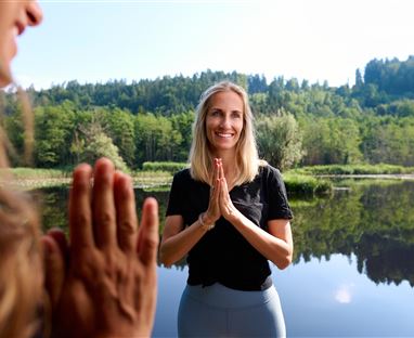 A woman stands by the shore of a tranquil body of water and smiles. In the background, green trees and a clear sky are visible.