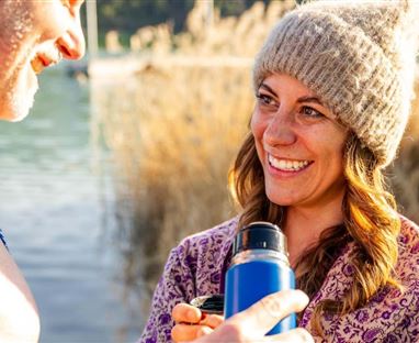 A cheerful couple by the water, laughing together. The woman holds a thermos and is wearing a hat.