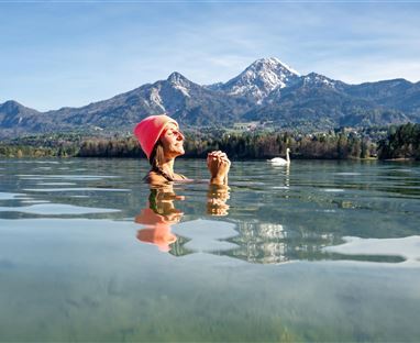 A person stands in the clear water of a lake with mountains in the background. The sky is blue and radiant.