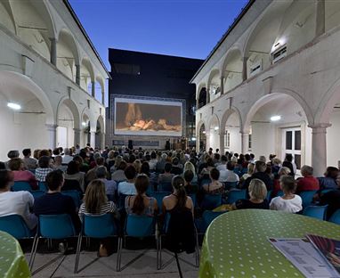A large crowd is sitting in a courtyard watching a movie on a screen. The sky is dark blue and the atmosphere is festive.