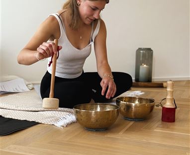 A person is sitting on the floor and playing with sound bowls. In the background, candles and a relaxing environment can be seen.