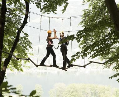 Zwei Personen stehen auf einer Hängebrücke zwischen den Bäumen und posieren fröhlich. Die Umgebung ist grün und naturnah, mit einem ruhigen See im Hintergrund.