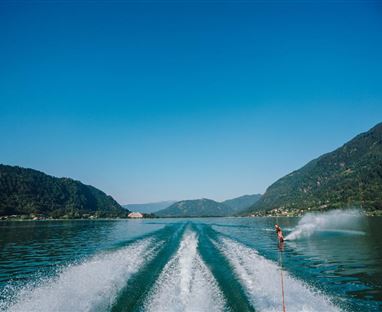 Un lago cristallino con onde leggere e un waterschier in azione. Sullo sfondo si vedono colline verdi e un cielo azzurro e splendente.