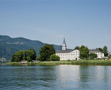 Eine ruhige Landschaft mit einem See und sanften Hügeln im Hintergrund. Im Vordergrund steht ein historisches Gebäude mit einem Kirchturm.