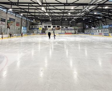 Eine Eishalle mit einer glatten Eisfläche und mehreren Eisläufern. Die Umgebung ist hell und gut beleuchtet, mit Werbebannern an den Wänden.