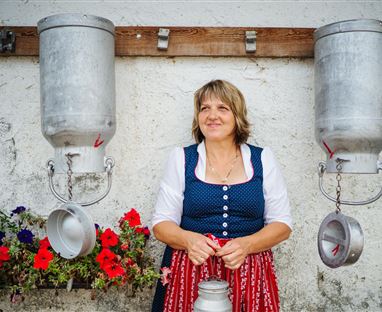 Eine lächelnde Frau in traditioneller Tracht steht zwischen zwei Metallbehältern. Im Hintergrund sind bunte Blumen zu sehen.