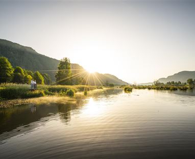 Ein ruhiger Fluss mit spiegelndem Wasser und grünen Ufern. Die Sonne geht hinter den Bergen auf und taucht die Landschaft in warmes Licht.