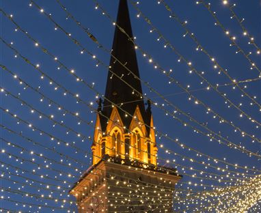 Ein schön beleuchteter Kirchturm vor einem nächtlichen Himmel. Lichterketten ziehen sich kunstvoll über den Platz.
