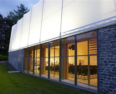 Un moderno edificio per eventi con grandi finestre e un interno ben illuminato. All'esterno, la facciata è in pietra e con una tela bianca.