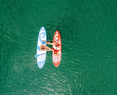 Zwei Personen paddeln auf Stand-Up-Paddle-Boards in einem klaren grünen Wasser. Eines der Boards ist blau und das andere rot.