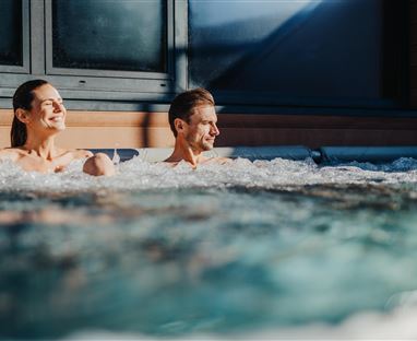 Ein Paar entspannt in einem Whirlpool. Die Sonne scheint und schafft eine gemütliche Atmosphäre.