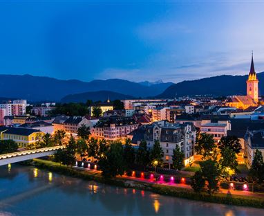 Eine malerische Stadtansicht bei Nacht mit beleuchteten Gebäuden am Fluss. Im Hintergrund ragt ein Kirchturm empor, umgeben von Bergen.