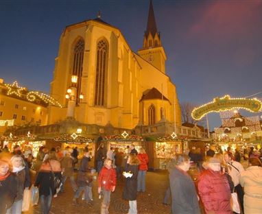 A festive Christmas market at dusk with many lights and cheerful people. In the background, a large church can be seen.