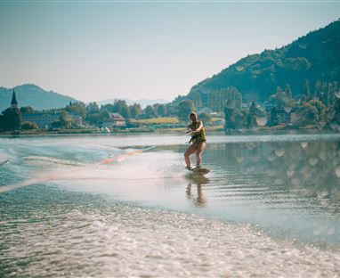 Eine Person fährt auf einem Wakeboard über ruhiges Wasser. Im Hintergrund sind Berge und eine kleine Stadt zu sehen.