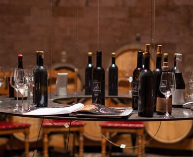 Una elegante enoteca con una varietà di bottiglie di vino e bicchieri su un tavolo. Sullo sfondo si vedono botti di vino e sedie rustiche.