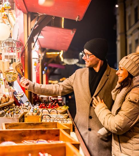 Ein Paar schaut sich die Waren an einem Weihnachtsmarkt an. Die Stände sind festlich beleuchtet und die Atmosphäre ist gemütlich.