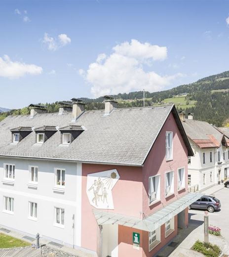 Ein malerisches Dorf mit bunten Häusern und einer Kirche im Hintergrund. Die Umgebung ist von grünen Hügeln und blauem Himmel geprägt.