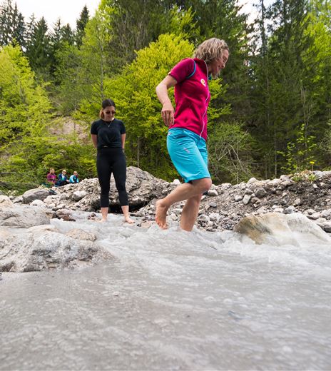 Two people are standing in a clear creek while balancing on the stones. In the background, lush green trees can be seen.