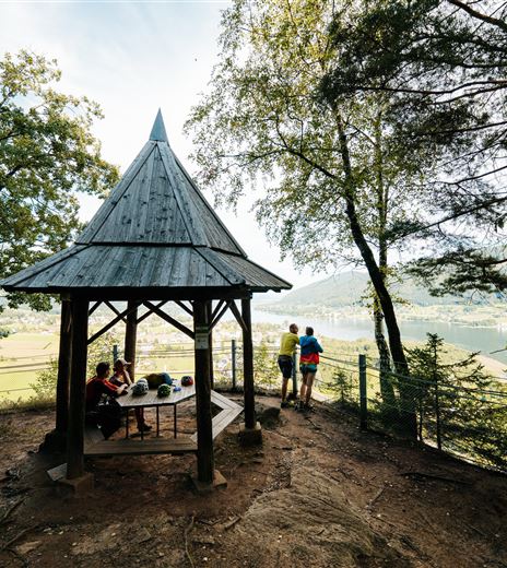 A wooden pavilion surrounded by trees with a view of the lake. Two people are standing and enjoying the scenery.