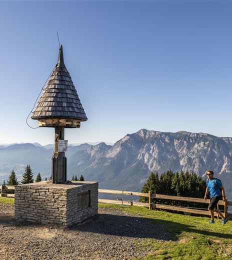Eine Wandergruppe auf einem Berg mit Blick auf majestätische Berge. Im Vordergrund steht ein kleiner Aussichtspunkt mit einem geschützten Dach.
