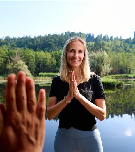 Una donna è in piedi sulla riva di un'acqua calma e sorride. Sullo sfondo si possono vedere alberi verdi e un cielo limpido.