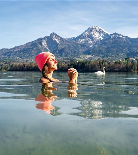 Una persona è in piedi nell'acqua limpida di un lago con montagne sullo sfondo. Il cielo è blu e radioso.