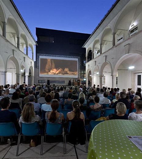 Eine große Menschenmenge sitzt in einem Innenhof und schaut einen Film auf einer Leinwand. Der Himmel ist dunkelblau und die Atmosphäre ist festlich.
