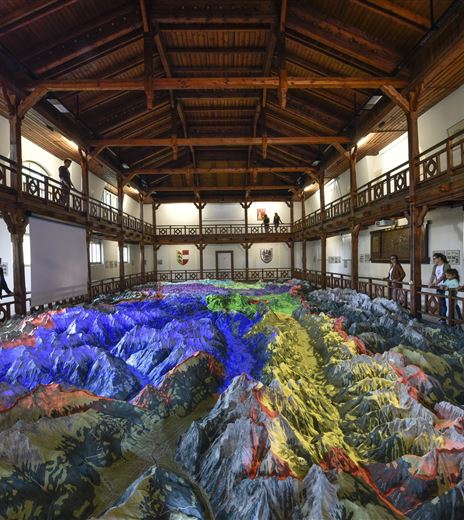 Un grande modello 3D di un paesaggio montano all'interno di un edificio storico. I visitatori esplorano la mappa topografica dettagliata con illuminazioni LED colorate.
