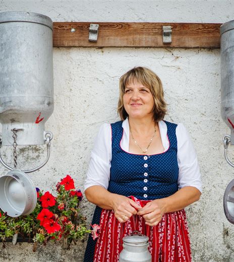 Eine lächelnde Frau in traditioneller Tracht steht zwischen zwei Metallbehältern. Im Hintergrund sind bunte Blumen zu sehen.