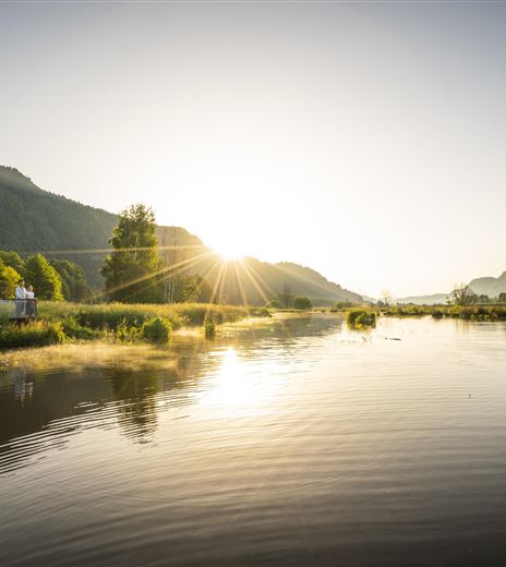 Un fiume tranquillo con acqua riflettente e sponde verdi. Il sole sorge dietro le montagne e tinge il paesaggio di luce calda.