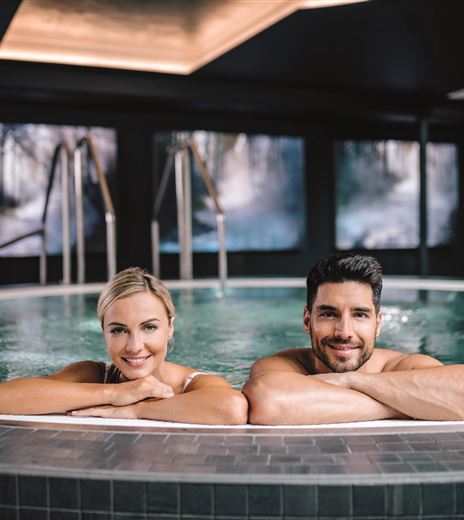 Ein Paar entspannt in einem modernen Whirlpool. Im Hintergrund sind elegante Bilder und eine gemütliche Atmosphäre zu sehen.