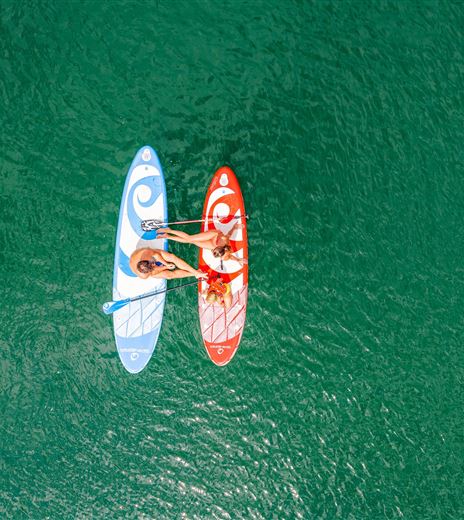 Due persone fanno paddle su tavole da paddle board in un'acqua verde chiara. Una delle tavole è blu e l'altra è rossa.