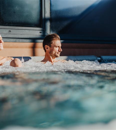 Ein Paar entspannt in einem Whirlpool. Die Sonne scheint und schafft eine gemütliche Atmosphäre.