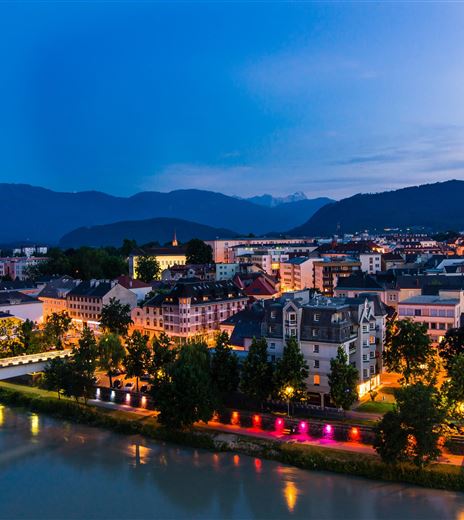 Eine malerische Stadtansicht bei Nacht mit beleuchteten Gebäuden am Fluss. Im Hintergrund ragt ein Kirchturm empor, umgeben von Bergen.