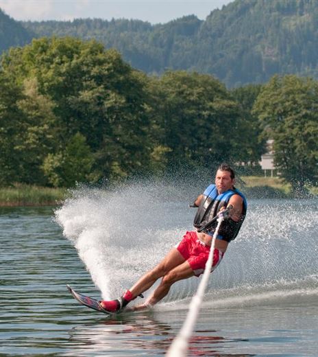 Ein Mann fährt Wasserski auf einem ruhigenSee. Im Hintergrund sind Bäume und eine Kirche zu sehen.