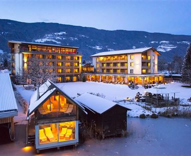 Un resort invernale con edifici illuminati nella neve. Sullo sfondo ci sono montagne e un lago tranquillo.